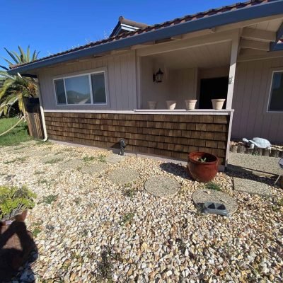 Charming cottage exterior with a wooden porch, potted plants, and a gravel pathway under a clear blu.
